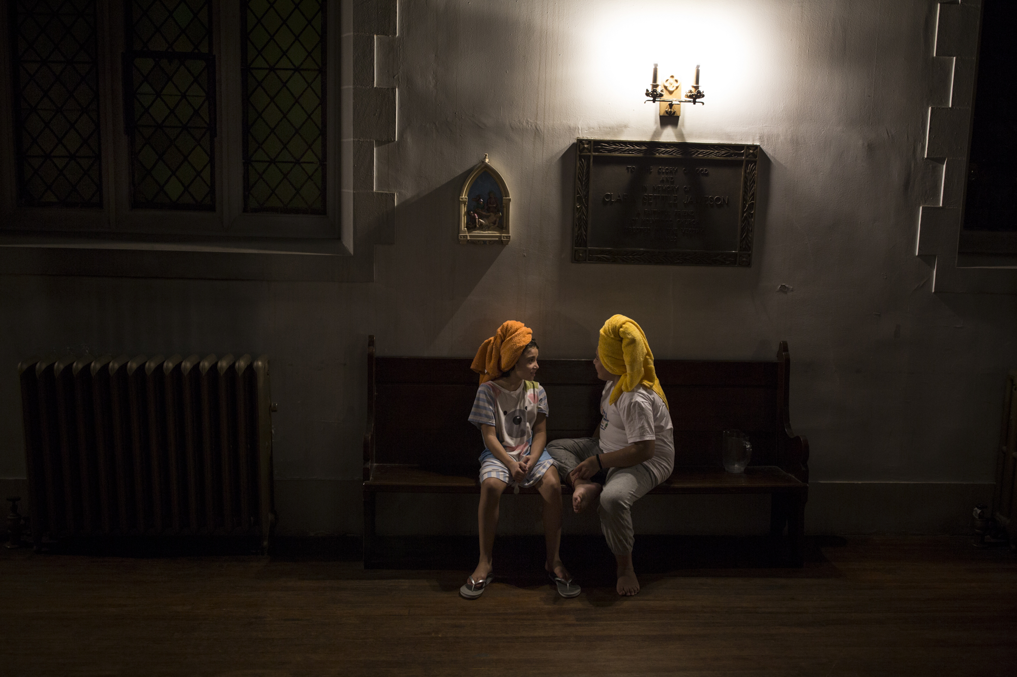 After taking a shower, Daniela, the one on the left, and her sister Dulce, are sitting down in one of the church's pews, waiting for their hair to dry. "We never thought of living in a church," says Dulce. ¨I feel there are ghosts all around me and they see me. But I also like living here. It is a beautiful place¨.
Since August 17, 2017, Dulce, Daniela and their brother, David, live, together with their mother, Amanda Morales, as refugees in the Episcopal Church of Holyrood, in Washington Heights, to avoid the deportation of their mother back to Guatemala.
For Dulce and Daniela, 10 and 9 years old respectively, to live in a church as refugees has not been easy, given that they had to leave behind their house, school and friends in Long Island. Their mother was the first undocumented migrant, in the last three decades, to take public sanctuary in the city of New York.
After they moved to the church, Dulce and Daniela have suffered episodes of anxiety for fear that their mother will be deported and separated from them and her brother Daniel, 3 years old.
This photo I took one of the many nights that I slept with Dulce, Daniela and her family in the church. After bathing Dulce and Daniela were playing in the sanctuary of the church and when I saw that they had sat on a church´s pew, I asked them if I could take a picture, and they said yes. At first they were posing for the picture and playing, but later they forgot I was there and that's when I captured this image. October 20th, 2017. Manhattan New York